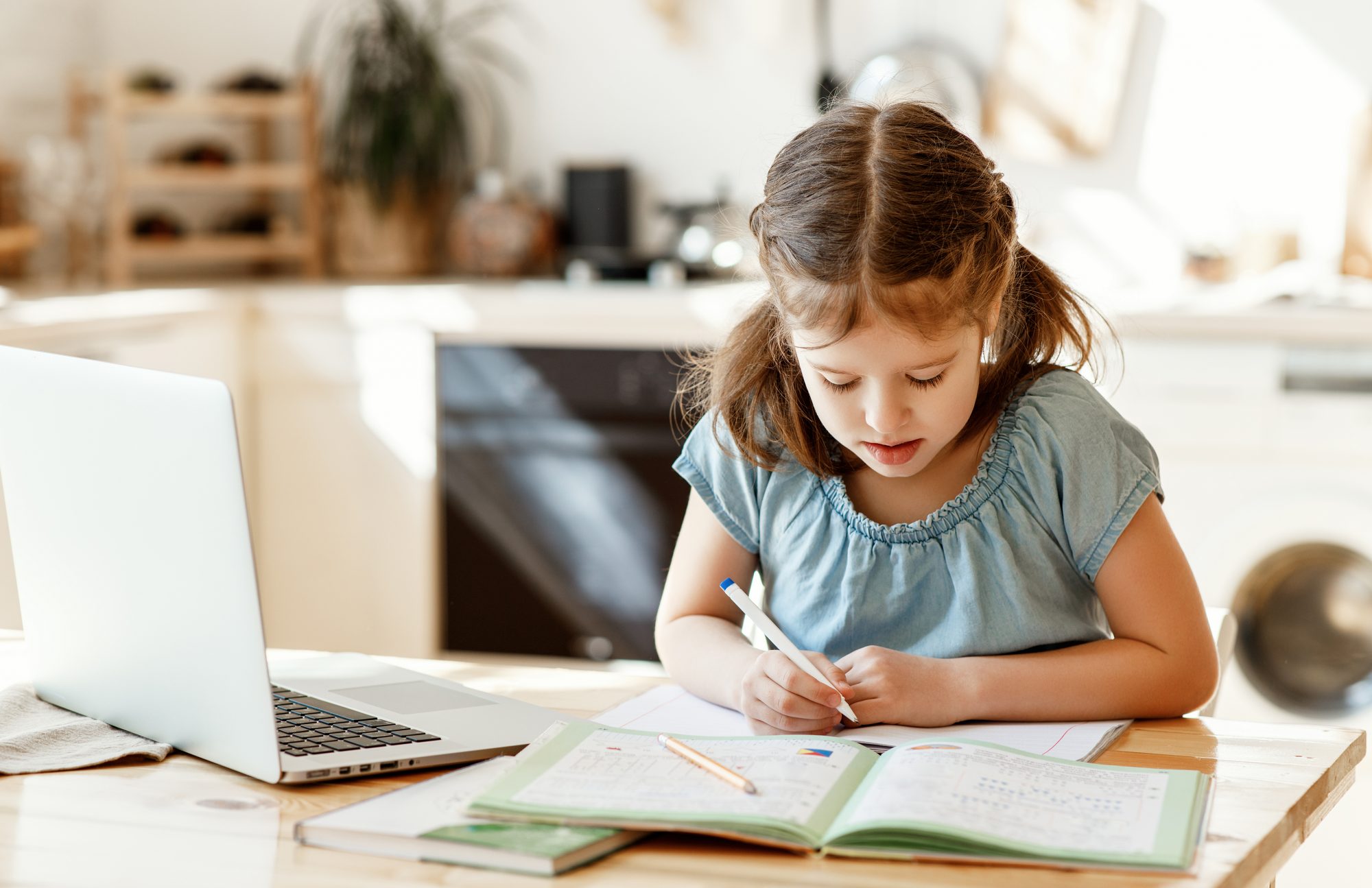 Little girl sitting at table doing homework with a laptop and a workbook