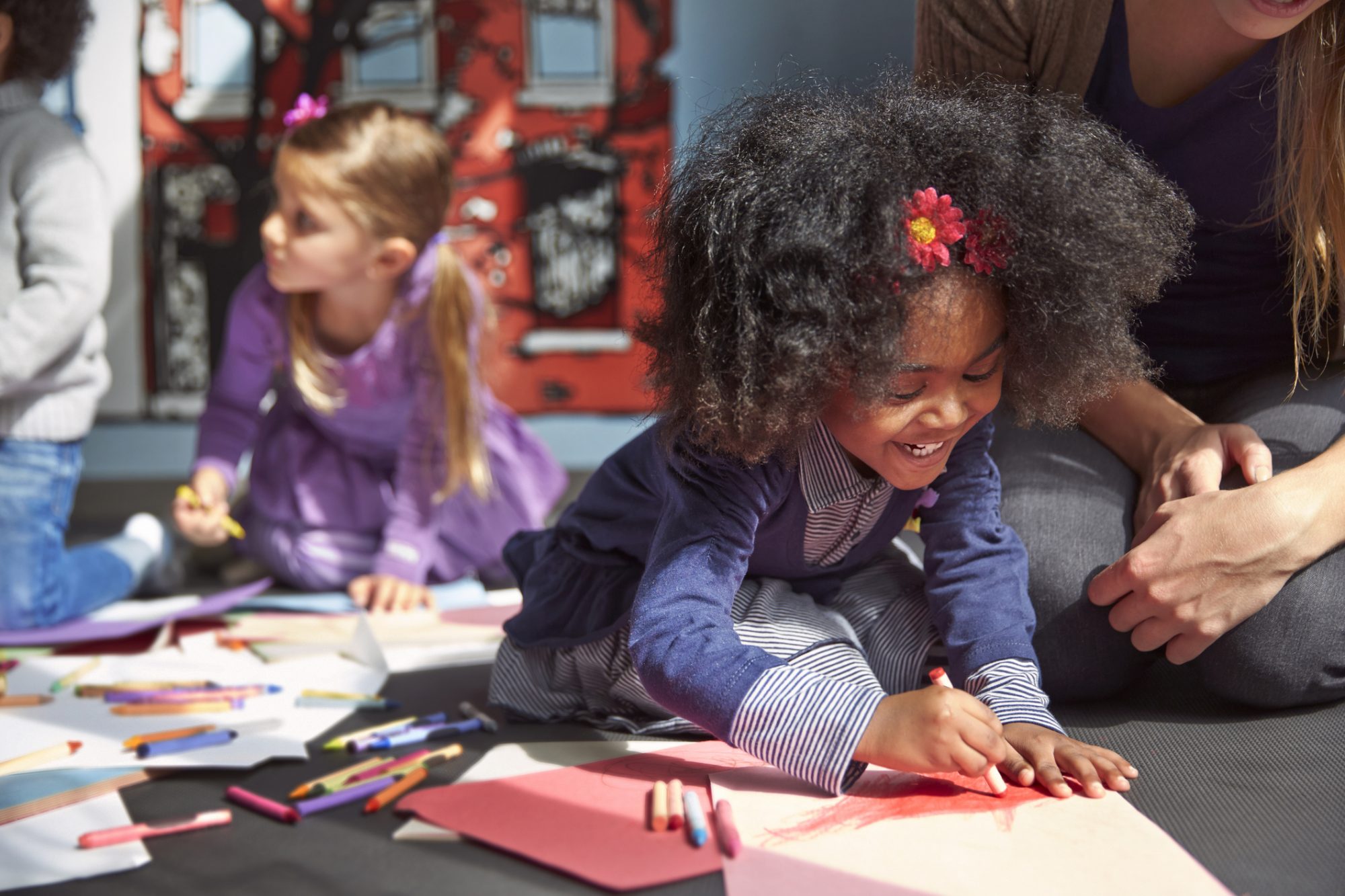 An image of a girl drawing on paper while sitting by her teacher in kindergarten.